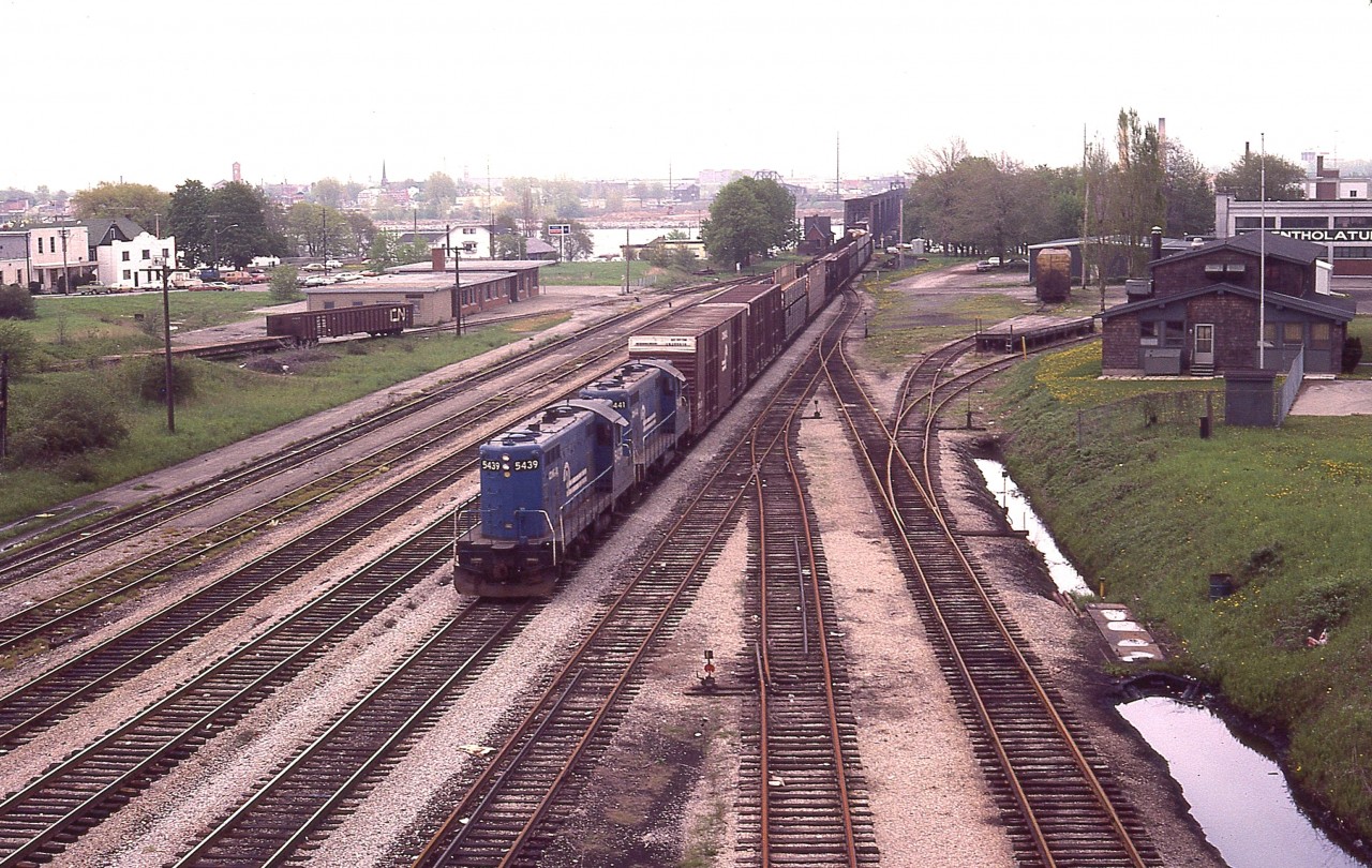 To clarify the location of the old CN Crew Hostel which I offered up the other day, here is a view of the eastern most part of CN Fort Erie trackage, taken 10 years before the Hostel closed.  It can be seen on the right; and the old MCRR/TH&B station on the left, with a Conrail transfer behind CR 5439 and 5441 just coming into Canada.  The USA is seen in the far background. Lots to look at here. In the background also, on the right, a yellowish rail car is parked on a stub. This was a Boeing car; one was spotted here on occasion due to the Fleet Aircraft company located nearby.
It is a rather gloomy day, and although this is not a factor, I scribbled in my notes that Mount St. Helens had blown up the previous day.  Just a historical note.
