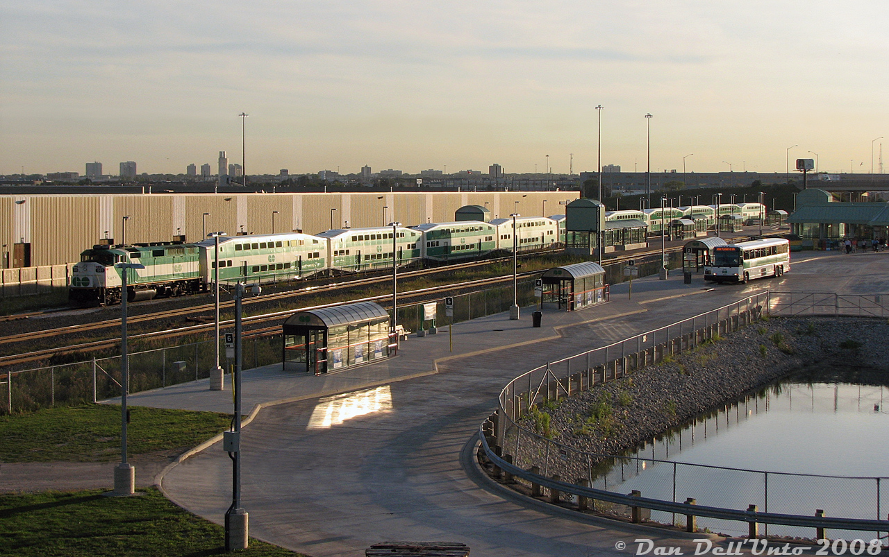 Evening "Train-Bus" at Bramalea:

Bramalea GO station was a much simpler place, pre-Metrolinxization. GO F59PH 561 sits on the Bramalea commuter lead, aka "Platform 3", after having just arrived with evening train #269 (Union-Bramalea), the last Georgetown train of the day.  A group of tired Brampton commuters can be seen making their way from the underground passage exit to one of the many GO D4500 commuter coaches (probably a 21-2200-series) operating on the "Mount Pleasant GO Station" connecting train-bus, for those wanting (daring?) to continue further into Brampton. The relatively new bus loop here was built in 2006 to accommodate the growing number of GO and Brampton Transit routes servicing the station, including GO's popular Highway 407 bus service to York University (always a hit with the local student residents). The main 1974-built station building is partially visible on the upper right.

From this vantage point on the Bramalea Road overpass, there's nothing really discernable in the "Brampton Skyline" except the long grey low roof of the nearby Ford parts distribution warehouse beyond the Steeles Avenue overpass (that once had rail service, but was all demolished in recent years). The building to the left was one of Kuehne & Nagel's distribution warehouses in the area (that still had its rail siding intact, but hadn't seen a boxcar at its roll-up doors in many years).

Everything here including station buildings, bus loop, platform and shelters, and stormwater pond was replaced and rebuilt starting in the late 2010's with a larger, more modern "Metrolinx style" station to serve the ever-growing Brampton population.
