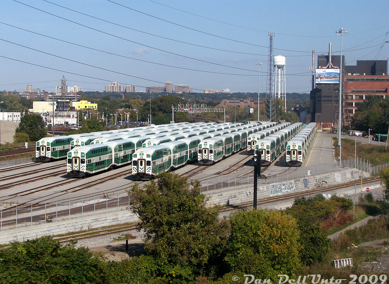 Afternoon layover at GO Transit's Don Yard east of Union Station shows multiple 10- and 12-car train consists awaiting their call to PM rush hour service in a few hours. Cab cars visible from left to right are 216, 202(?), 2??, 236, 206(?), 250, 209, 251, 204, and 237, with various MP40 and F59 units on the other ends.

Don Yard used to be an old CN freight yard for the longest time, dating back to the steam era, until it was taken over by GO Transit around 2006-07 and converted to store GO trains on layover (like Bathurst North Yard years earlier). The track running along the bottom of the retaining wall was the spur from the Union Station Rail Corridor to the Toronto Harbour Commission trackage in the Toronto portlands (Eastern Harbour Terminals) including Keating Yard at the foot of the Don Roadway, the Toronto Port Authority, the Ashbridges Bay Wastewater Treatment Plant, and to the old Wilson Yard (a small storage yard just south of Don Yard). All of that trackage has since been removed after rail service to the portlands was discontinued (last run in 2018).

The CN Bala Sub can be seen curving up the Don Valley to the north off the USRC (above the leftmost cab car), and the CN Kingston Sub curving off in the distance beyond the signal bridge. The large building on the right with the water tower is the old Lever Brothers/Unilever/Korex soap plant (once had rail service, but recently closed for good in August 2009 after the company declared bankruptcy during to a labour disruption that started in 2008). The two old red buildings beyond that are from the old Consumers Gas Station B coal gas manufacturing facility that was once around Eastern Ave & Booth (they once had multiple sidings and rail service too). Some apartments in East York are visible in the distance above the foliage canopy  (possibly around Victoria Park/Dawes Rd).

This was a rather difficult shot to get at the time, as in 2009 we weren't blessed with the abundance of cheap commercially available drones, there were no condos, bridges or overpasses nearby to shoot from, stopping here is not advised, and you couldn't exactly walk up the side of the Gardiner Expressway to this spot without quickly being branded a looney and getting a wellness visit from a few Crown Vics ("Yes officer, I'll come down peacefully, I just need to shoot the GO trains in the train yard first"). The side barriers and guardrails also blocked this view from most cars. But a ride bouncing around at the back of a GO D4500 commuter coach on the Barrie-King City-Vaughan run (that only went down the Don Valley on certain days, at certain times, depending on certain operators) gave just enough elevation to snap a photo looking backwards and get all of Don Yard in view. I remember sending it to the late Ray Kennedy (https://trainweb.us/oldtimetrains/TTR/aerial_2009.htm) at the time, and he seemed quite impressed I was able to get a shot of the new GO yard from this angle.