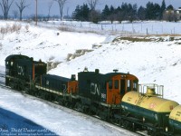 CN S13 units 8611 and 8621 bracket slug B7 on a hump set, working the <a href=http://www.railpictures.ca/?attachment_id=51354><b>Toronto Yard</b></a> (later renamed MacMillan) <a href=https://www.railpictures.ca/?attachment_id=57709><b>hump</b></a> as they sit on the pullback track north of Rutherford Road overpass. Two fresh-looking Dupont Canada anti-knock compound tank cars are coupled near the head end.<br><br>When the yard opened in the mid-1960's, they used <a href=http://www.railpictures.ca/?attachment_id=38976><b>S13-slug-S13 sets</b></a> designated in an H1-H2-H3 configuration (note red lettering above roadnumbers), where H1 was the main unit outfitted with hump control, radio and signal equipment, H2 was the slug (converted from <a href=https://cptdb.ca/wiki/index.php/Canadian_National_Railway_300-314><b>retired S3 units</b></a>) and H3 was a trail-only unit with no special equipment onboard. The <a href=https://cptdb.ca/wiki/index.php/Canadian_National_Railway_8600-8625><b>8600's</b></a> were a slightly heavier duty specified MLW S13 than the <a href=https://cptdb.ca/wiki/index.php/Canadian_National_Railway_8500-8522><b>8500's</b></a>, equipped with GE 752 traction motors, full automatic transition, MU, 26L brakes and extra weight. This power was later displaced by more powerful modified GP38-2 units and new GMD slugs in the late 70's.<br><br>This area of Vaughan (the old hamlet of Maple/Sherwood) was still very rural at the time, with no Canada's Wonderland or housing developments visible north of the yard. The farm and old stone farmhouse at the upper right was about where Seafield Road is today (suburban neighbourhood built in the 90's), and the farm and barns in the distance were way up at Major Mackenzie and Jane Street (southwest corner, where the Fortinos plaza is today).<br><br><i>Original photographer unknown (possibly Robbin Rekiel), Dan Dell'Unto collection slide.</i>