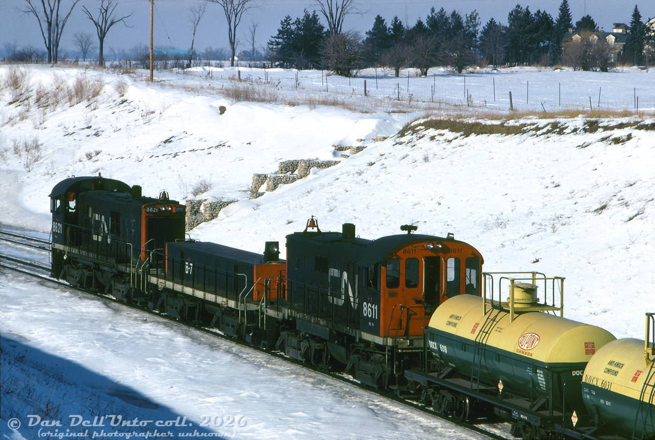 CN S13 units 8611 and 8621 bracket slug B7 on a hump set, working the Toronto Yard (later renamed MacMillan) hump on the pullback track north of Rutherford Road overpass. Two fresh-looking Dupont Canada anti-knock compound tank cars are coupled near the head end. When the yard opened, they used S13-slug-S13 sets designated in an H1-H2-H3 configuration, where H1 was the main unit outfitted with hump control, radio and signal equipment, H2 was the slug (converted from retired S3 units) and H3 was a trail-only unit with no special equipment aboard. The 8600's were a slightly heavier duty specified S13 than the 8500's, equipped with GE 752 traction motors, full automatic transition, MU, 26L brakes and extra weight.

This area of Vaughan (the old hamlet of Maple/Sherwood) was still very rural at the time, with no Canada's Wonderland or housing developments visible north of the yard. The farm and old stone farmhouse at the upper right was about where Seafield Road is today (suburban neighbourhood built in the 90's), and the farm and barns in the distance were way up at Major Mackenzie and Jane Street (southwest corner, where the Fortinos plaza is today).

Original photographer unknown (possibly Robbin Rekiel), Dan Dell'Unto collection slide.