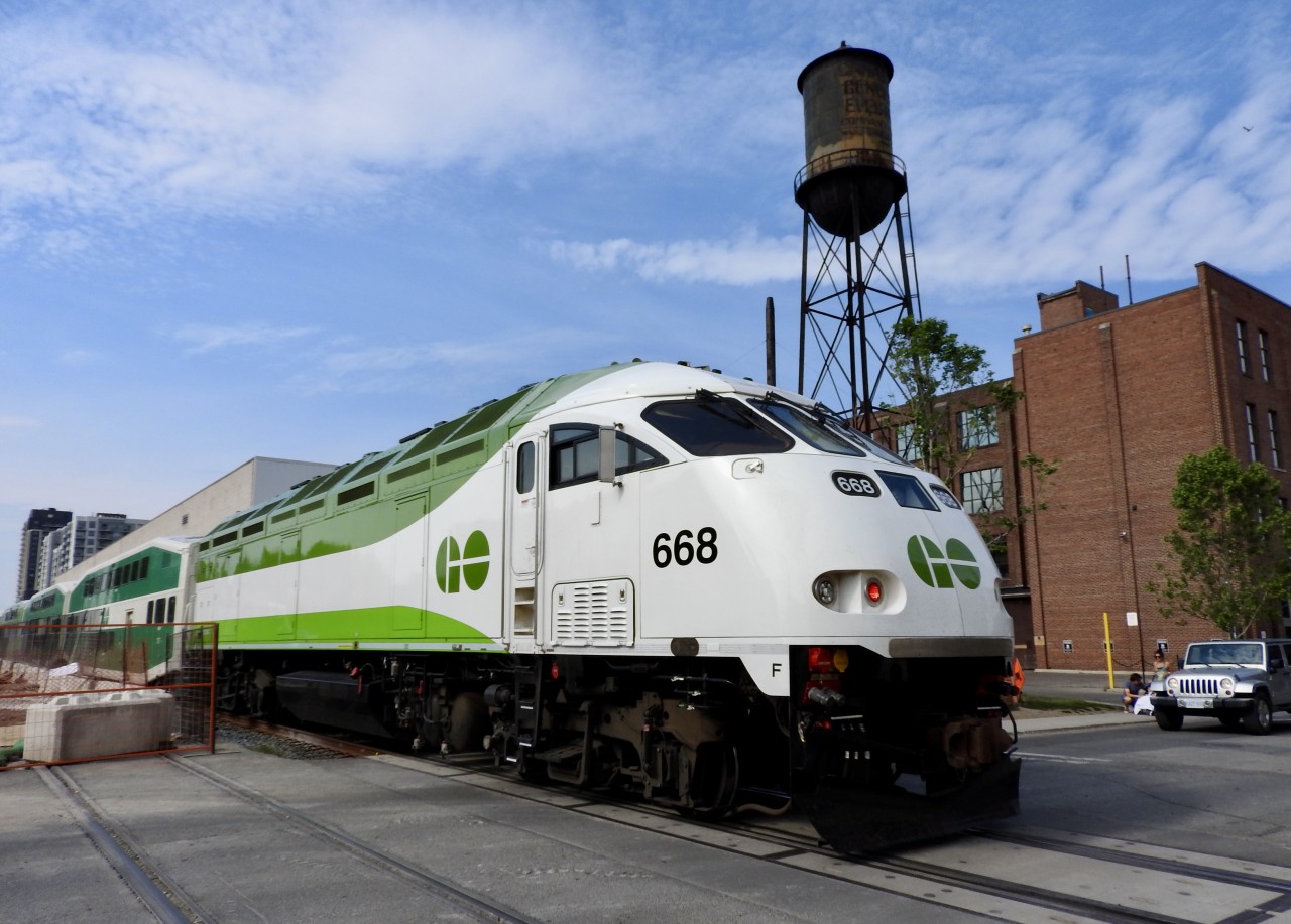 6/02/21 GO MP54AC #668 is shoving hard on its consists as it heads up the Newmarket Sub towards Barrie, Ont. The locomotive is just passing Wallace Ave, in The Junction area of Toronto. The big water tower in the background has an interesting history.

Canadian General Electric (CGE) Ward Street Works plant in Toronto's Junction-Wallace Emerson neighbourhood, which is famous for its towering wooden water tower. 

The factory, built in the early 1920s at 224 Wallace Avenue, once manufactured lamp bases and electrical supplies. It has since been transformed into vibrant creative, studio, and commercial spaces. The old plant even served as the former home of video game developer Ubisoft. 

The CGE complex is best known for a few distinct, prominent local landmarks:
* The Wooden Water Tower: It is one of the very last original wooden water towers still standing in the city. The City of Toronto gave it heritage status in 1993.

* The Ghost Sign: Painted high atop the water tower is a faded, historic sign that reads, "Canadian General Electric Company Limited - Ward Street Works." 

This photo will never be seen again due to the Metrolinx expansion of the Newmarket Sub. If you visit today, they have installed a second track. Sarting at Bloor St the double track rises up to a grade that eliminated the very busy CP/CN diamond at West Toronto. The times, they are a changin