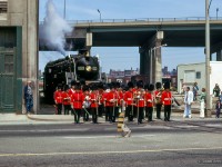 With full pomp and circumstance provided by the Band of the Royal Regiment of Canada, CNR 6060 is paraded towards Toronto's harbourfront at the Lakeshore Boulevard crossing, just east of Bathurst Street (note the<a href=https://www.railpictures.ca/?attachment_id=30123> tower at Bathurst north yard</a> in the distance.)  It is Victoria Day weekend, and opening day for the Canadian Railway Historical Association's Toronto & York Division Harbourfront Railway Museum, just to the south of Lakeshore Blvd.  6060 is taking part by hauling museum equipment to the new site, in this image <a href=https://www.trainweb.us/oldtimetrains/photos/cnr_diesel/7988_P.jpg>pulling MLW S2 7988.</a>  The museum would close and equipment dispersed into the mid 80's.  At left is the old 1928 Loblaws warehouse, the facade still standing today as part of the West Block condo development.


<br><br><i>Scan and editing by Jacob Patterson.</i>