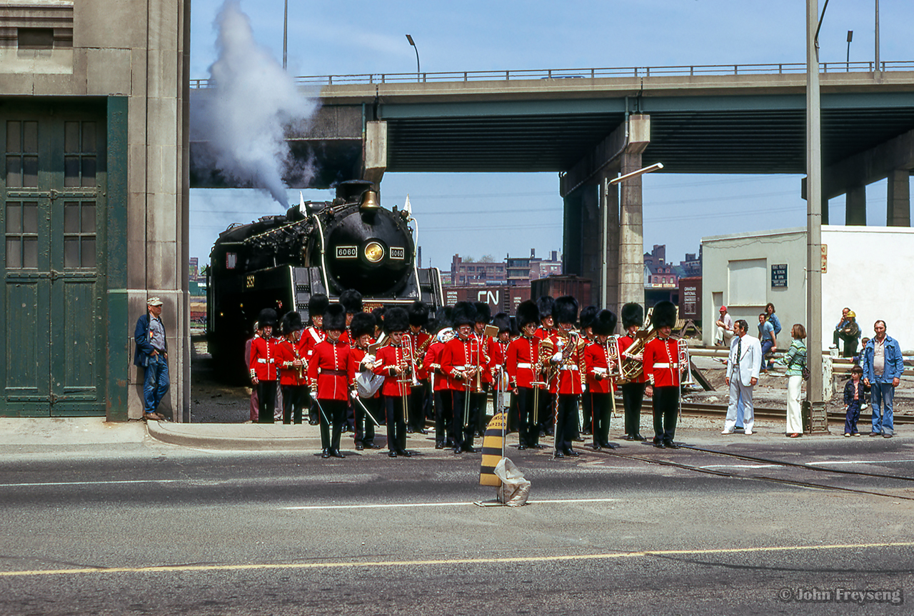With full pomp and circumstance provided by the Band of the Royal Regiment of Canada, CNR 6060 is paraded towards Toronto's harbourfront at the Lakeshore Boulevard crossing, just east of Bathurst Street (note the tower at Bathurst north yard in the distance.)  It is Victoria Day weekend, and opening day for the Canadian Railway Historical Association's Toronto & York Division Harbourfront Railway Museum, just to the south of Lakeshore Blvd.  6060 is taking part by hauling museum equipment to the new site, in this image pulling MLW S2 7988.  The museum would close and equipment dispersed into the mid 80's.  At left is the old 1928 Loblaws warehouse, the facade still standing today as part of the West Block condo development.


Scan and editing by Jacob Patterson.