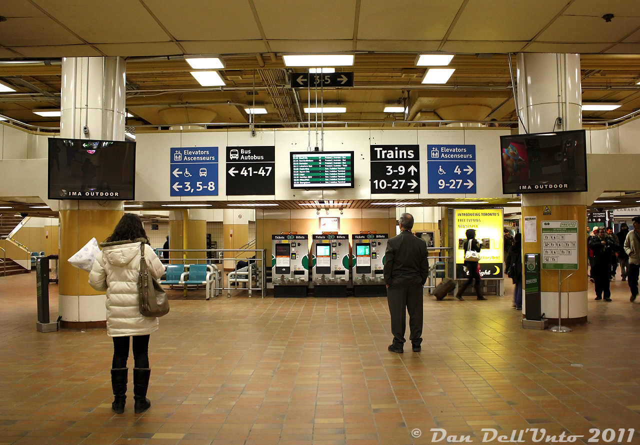 Everyone's favourite concourse: the GO concourse, in the basement of Toronto Union Station. A daily visit for many, the bane of their existence for some. Often smelly, crowded, noisy from trains rumbling above, frequented by various solicitors, occasionally flooded ("Man the GO Ark!") and more often than not full of the intoxicating smell of Cinnabons behind pumped out nearby.  It's quieter down here tonight as evening commuters keenly watch the departure boards for the platform number of their GO train or bus trip to appear. Once some unknown higher power posts that magic number, it's off to the races through the maze of overhead walkways and staircases to get to the correct platform to board in time (don't forget to stamp your 10-ride ticket or tap your Presto first, they don't like it when you forget). The "t.o.night" being advertised by the ticket machines was a briefly popular evening commuter newspaper at the time, with a basic glazing over of local/national news, pop culture and current events mixed in with ads, games/puzzles, horoscopes and a somewhat amusing commuter shoutout section (I think it lasted until 2014-ish). Signage advises commuters, many set in their paper ticket ways, of the benefits and usage of the new Presto farecard system that the GO and Accenture staff at 130 Adelaide spent much time developing.  The site of the original GO Transit commuter concourse was the present VIA arrivals area under the Great Hall (which can be seen in the 1976 movie Silver Streak, thinly disguised for AM Road, complete with GO's ticket gates for pre-Proof-Of-Payment days). GO moved into this new concourse in 1979, build in the basement in the east end of Union Station that was once a postal handling facility. There were ticket gates here too (that can be seen in NFB's "After the Axe" film) until they were removed when GO changed to the POP system in the mid-late 80's.  The GO concourse was a time capsule in itself, featuring 1970's yellow, orange and brown earth tones, no shortage of round pillars supporting the trains rumbling just above the ceiling structure, and elevated walkway maze leading to various track platform stairways. Food vendors and aromas were in abundance, from the likes of McDonalds, Mr. Sub, Mmmuffins (Mmmmmmmmm), Dairy Queen, Mr. Sub and of course Cinnabon. The look and feel of the GO concourse remained largely unchanged until it was closed in mid-August 2015. GO passengers shifted to a new York Concourse, in order for the Bay Concourse to get a complete gutting and modernization.  The retro Toronto nostalgia buff types and social influencers on the 'grams would have have a field day down here if this relic in time still existed, alas, they'll have to get their kicks over at Spadina subway station or Dupont instead.