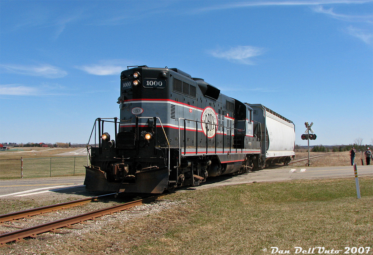 If you opened a dictionary and looked up "Shortline Railroading", this photo would sum it up nicely: the Orangeville-Brampton Railway's twice weekly run to Streetsville to interchange traffic with CP (and to work the few industries enroute) has just left the suburban chaos of Brampton and it making its way north through the Caledon countryside. Traffic today consists of just a single covered hopper, and with interchange work done it's a straight shot all the way back to Orangeville.

Cando Contracting (CCGX) 1000, an old 1957 GMD GP9 (ex-QNS&L 157) that was the lone assigned power for the OBRY, is running long-hood-forward and laying on a little bit extra horn for the three photographers giving chase as it cruises past the Brampton-Caledon Airport and over McLaughlin Road, near the hamlet of Victoria. There's actually four RP.ca photographers here: Dave Young and Mike Da Costa shooting at the crossing, me behind the camera, and Steve Bradley in the cab as conductor on this run. Engineer Wayne was giving 1000's half-clogged Leslie Supertyphon S3J horn a good workout at each crossing on the trip north, and both were nice enough to invite us to ride around in the cab in Orangeville before the unit was put away for the day. A fun time was had by all, with a good amount of photos and memories to look back through.
