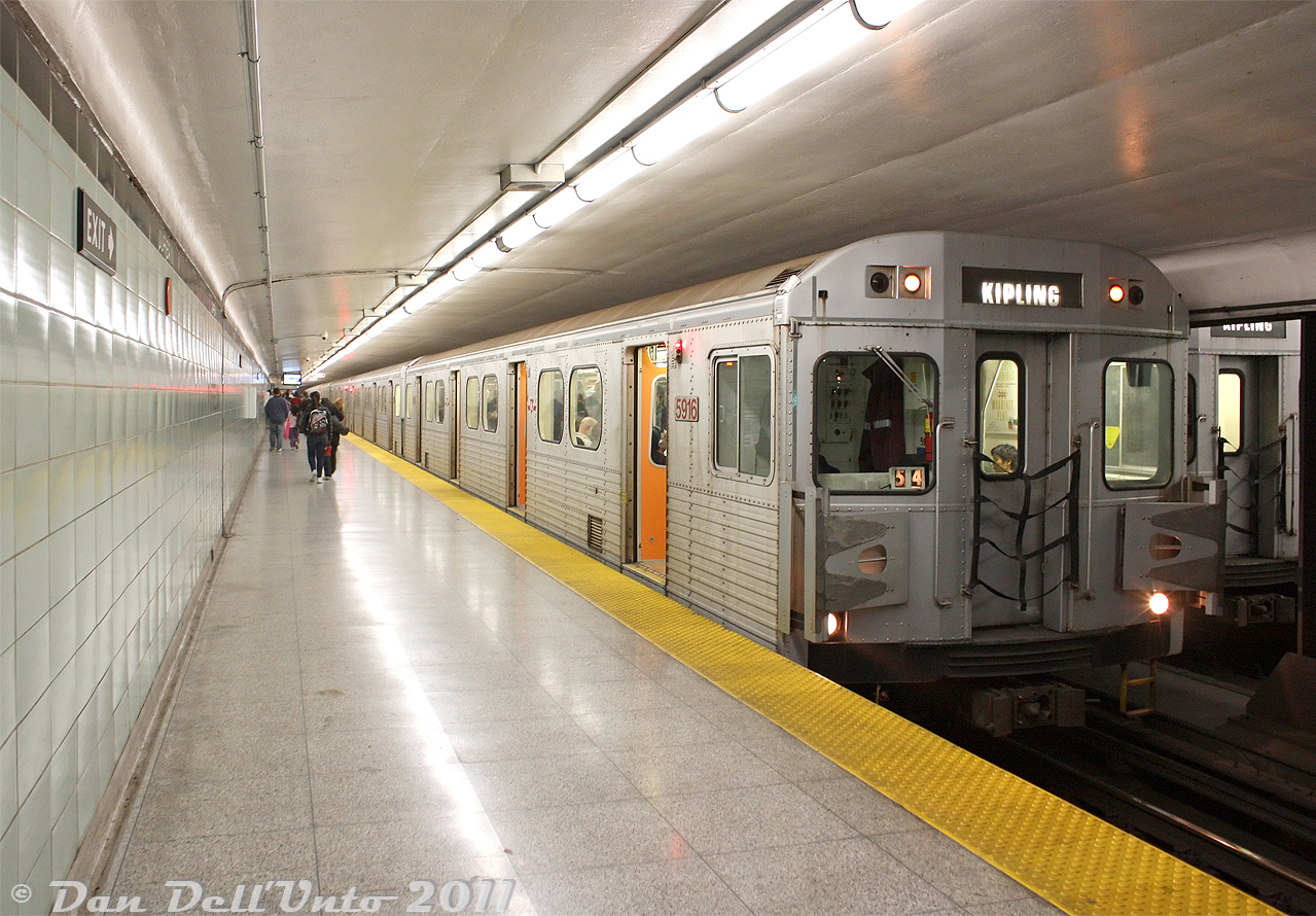 Operating on a westbound Bloor-Danforth run during rush hour, TTC 5916 leads a train of UTDC/Can-Car H6 subway cars westbound on the Bloor-Danforth line during a brief stop at Dufferin Subway Station. The open doors show off the distinctive orange doorways featured on the H6's. Another set of H6 subway cars sits on the adjacent platform, making their stop before continuing eastbound for Kennedy.