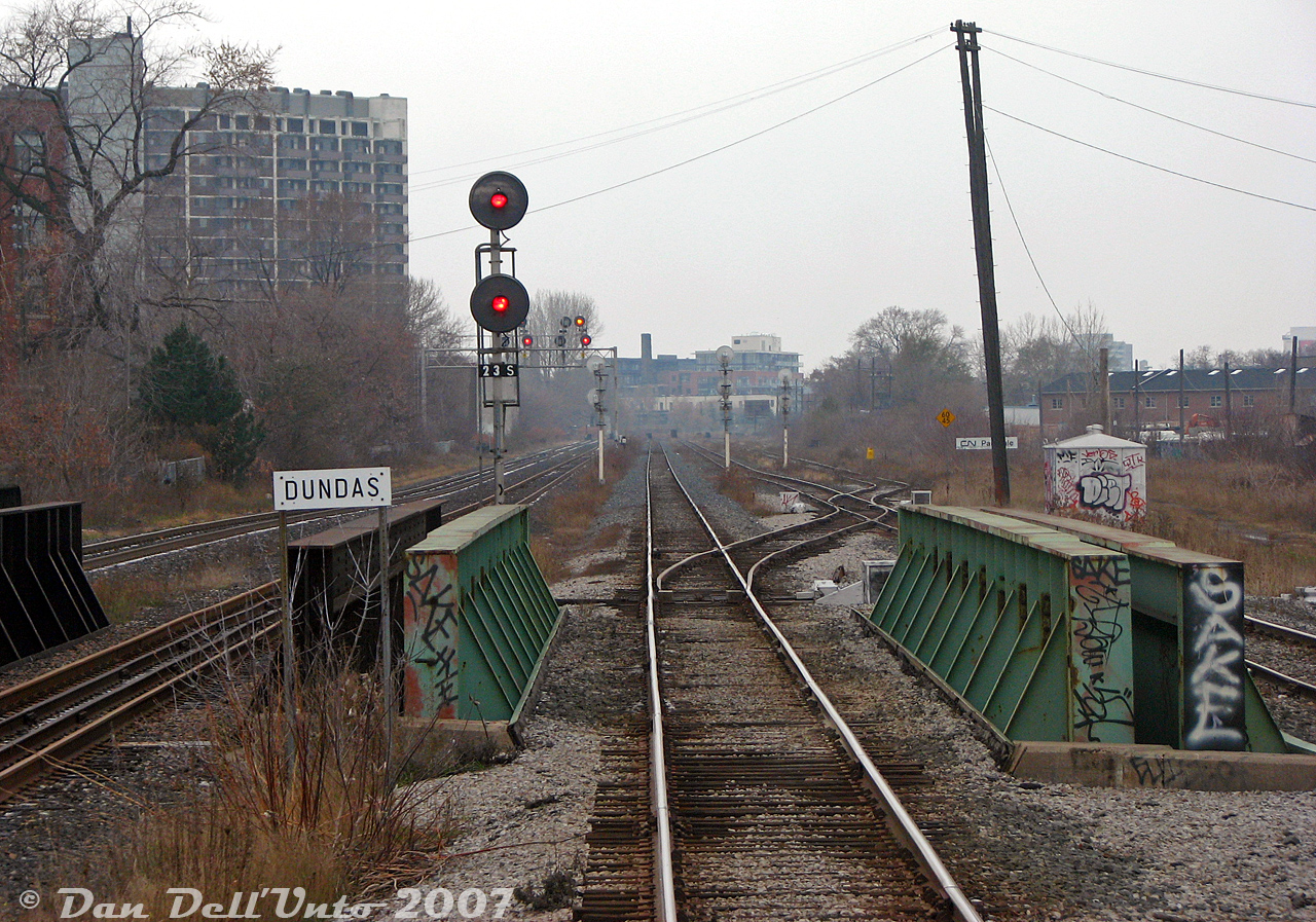 Another shot from the best seat on the train: riding in the back of GO Transit cab car 201 on late morning train #252 (Georgetown line, Bramalea-Union), we've just rattled over the crossover switch between CN's Weston Sub mainline tracks and over the steel girder bridge span over busy Queen Street West at CN Parkdale, click-clacking down the stick rail on the home stretch to Toronto Union Station downtown. We're on the south mainline track, with the north mainline visible to the right, along with the start of the CN Newmarket Sub (GO Barrie-line trains) branching off at the switch by the sign. The "last minute" crossover track here let Barrie-line GO trains use the south track of the Weston Sub to/from Parkdale. On the left are CP's two Galt Sub mainline tracks (GO Milton line), that at the time were completely separate all the way to West Toronto and weren't connected with the Weston Sub at all. This location has the mileboard for "CP Dundas" (after Dundas Street, further north at Mile 3.5 CP Galt Sub) however this location itself was still known as CP Parkdale in official timetables (Mile 2.3), but with nothing to really here of importance to note anymore. There was a really old wooden relic of a CP Parkdale sign that used to be around Lansdowne Ave in the distance, with a modern sign lazily nailed over it. Hopefully it went to a good home when Metrolinx removed it.

How the mighty have fallen: this part of Toronto was once home to not one but two decent-sized railway stations (CN and CP Parkdale, both demolished), small and medium-sized yards and associated facilities for both railways (all removed), and lots of industrial sidings and spurs for local factories and industries around Parkdale and Liberty Village (all gone). Parkdale railway-wise in 2007 had been reduced to just an interlocking plant where the Newmarket Sub (Mile 2.4) split off from the Weston Sub (Mile 2.5).

Around 2008, the (Metrolinx/CN) Parkdale interlocking was moved north closer to Lansdowne underpass to facilitate rebuilding the underpasses and Duffern Street, in order to eliminate the "Dufferin Jog" at Queen Street (continuing Dufferin Street below the rail bridges instead of its previous disjointed alignment). New modern signals went in replacing the old ones, some track realignments took place around this time with the unused spans to keep rail traffic flowing as the bridge spans were rebuilt or reconditioned, and several new spans added. More recent track diagrams now show Metrolinx has a total of 8 bridge spans here: 4 for active mainline tracks (x1 Galt Sub, x3 Weston Sub) as well as 4 extra spans for future track additions as-needed (x2 for future planned extensions of the Newmarket Sub, x2 for adding extra Weston or Galt Sub mainline tracks back).