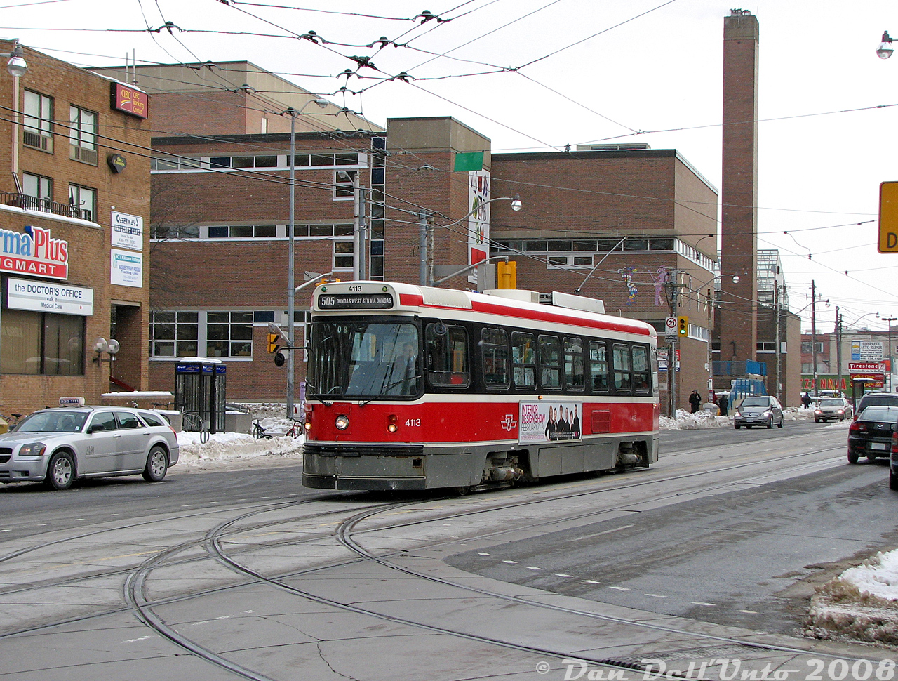 TTC CLRV 4113 has just passed the busy intersection of Dundas and Bloor and is about to swing into Dundas West Subway Station on one of the tracks in the foreground, finishing up a trip across Dundas Street from Broadview Station in the east end. Part of The Crossways Mall can be seen on the left, and in the background the old Bishop Marrocco Catholic Secondary School, built in the mid-late 1960's on the site of some commercial buildings and the small CPR "Bloor Street Yard" once located here just off the Galt Sub. The small commercial plaza south of there was at the time home to a rather shabby looking Zellers (originally Towers), Loblaws, and Coffee Time, among other stores.  More around Bloor & Dundas: Bloor GO Station: https://www.railpictures.ca/?attachment_id=59667 NW corner in 1965: http://www.railpictures.ca/?attachment_id=56302 NW corner 1989: http://www.railpictures.ca/?attachment_id=53600 SW corner 1965 http://www.railpictures.ca/?attachment_id=36187 Slightly further south http://www.railpictures.ca/?attachment_id=31303 Dundas West station, brand new: http://www.railpictures.ca/?attachment_id=51391 Departing Dundas West station: http://www.railpictures.ca/?attachment_id=51570