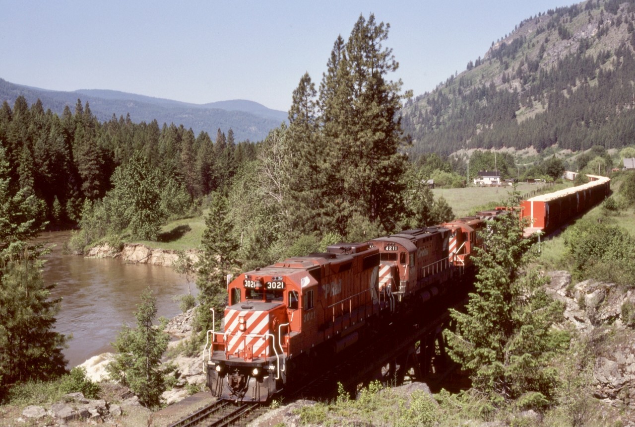 Eastward woodchip loads from Midway and Grand Forks sawmills bound for Kraft pulp mill near Castlegar dominate on an extra east with CP 3021 + 4211 + 8816 + 5023 (GP38-2, C-424, GP9, and GP35) crossing the Kettle River at mileage 82.7 approaching Cascade on Saturday 1984-06-16.  Another 1.6 miles east is another (and much bigger) bridge, the fifth of total five over the Kettle River, then the beginning of a significant ascent to the summit at Paulson Gap and Farron.

Today, this is the Columbia and Western Rail Trail.