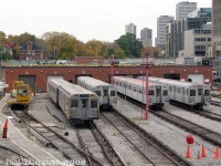 A nice selection of TTC subway equipment is pictured lined up outside the shops building at Davisville Yard, all typical cars serviced at Davisville at the time. From left to right are TTC flatcar (work car) RT-29, TTC H4 work cars RT-65 and RT-64, two sets of H5 subway cars, and a set of T1's. On the far right, the front car of a train of yet H5 cars sits on the third platform at Davisville Subway Station at Yonge and Davisville (with the midtown Toronto skyline along Yonge near Davisville-Eglinton visible in the distance).<br><br>Davisville Yard was the TTC's <a href=http://www.railpictures.ca/?attachment_id=41560><b>original 1950's maintenance base</b></a> for the newly opened Yonge Subway line, but by this point in time shared Yonge-University-Spadina line equipment with the larger and more modern <a href=http://www.railpictures.ca/?attachment_id=37314><b>Wilson Yard</b></a>. The H5 cars were strictly operated on the YUS, while the T1's handled the YUS and Sheppard Subway duties. The other two remaining revenue classes (<a href=http://www.railpictures.ca/?attachment_id=31197><b>H4</b></a> and <a href=http://www.railpictures.ca/?attachment_id=25985><b>H6</b></a>) were assigned to <a href=http://www.railpictures.ca/?attachment_id=36389><b>Greenwood Yard</b></a> for Bloor-Danforth line service with the older T1's.<br><br>There were two rail attractions to visit in this area of Toronto: the TTC Davisville Yards from this vantage point off the Kay Gardner Beltline Trail (the old <a href=http://www.railpictures.ca/?attachment_id=32616><b>CN Belt Line Branch</b></a> off the Newmarket Sub, that the city purchased in the early 1970's and turned into a popular walking trail) and the original <a href=http://www.railpictures.ca/?attachment_id=39041><b>George's Trains</b></a> nearby, along <a href=http://www.railpictures.ca/?attachment_id=52945><b>Mount Pleasant near Davisville</b></a>.
