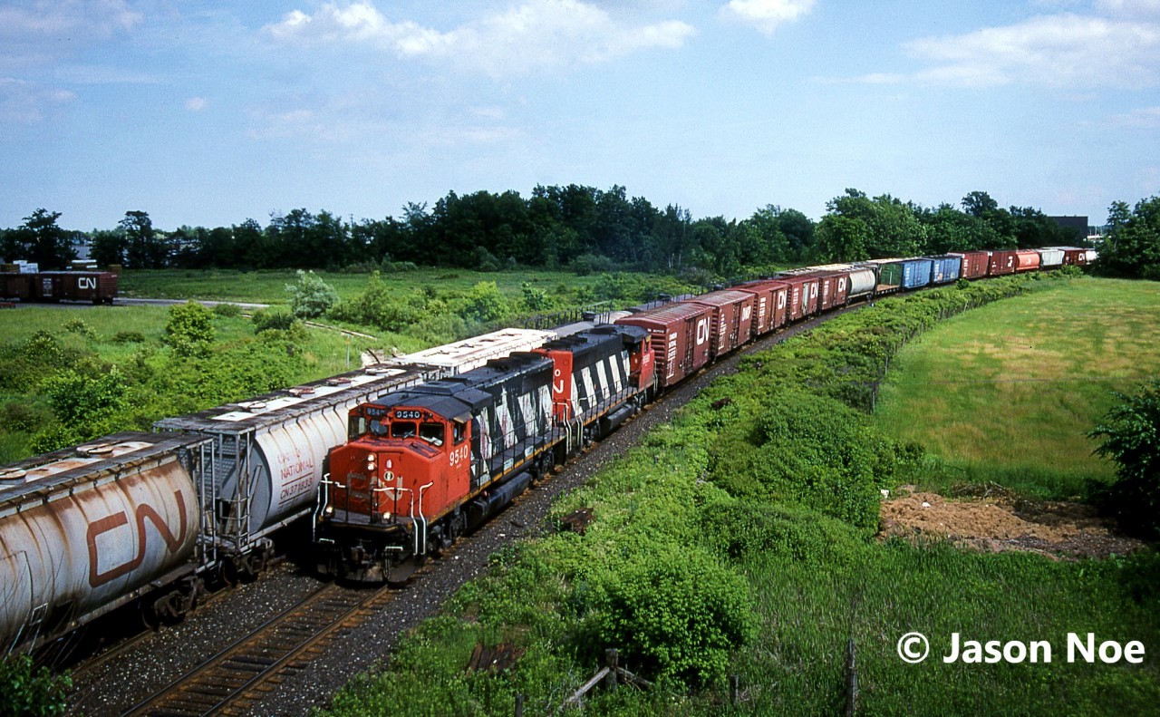 An eastbound CN train is viewed about to duck under Highway 401 with GP40-2L(W)’s 9540 and 9560 while meeting 363 at Milbase, which is located in Milton, Ontario on CN’s Halton Subdivision.