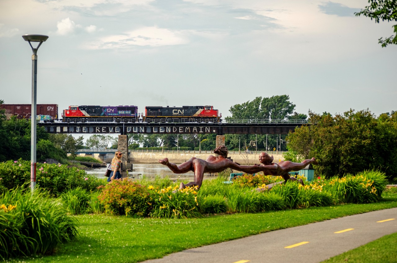 CN 402 crosses the bridge over the Rimouski river with SD75IACC #8357 in trail, a locomotive specially painted for suicide prevention. In the foreground is the sculpture titled La Rencontre Sous-marine, by local artist Roger Langevin. The bridge over the river was first built by the Intercolonial railway in the early 1870s, the girders were later replaced in the early 20th century.