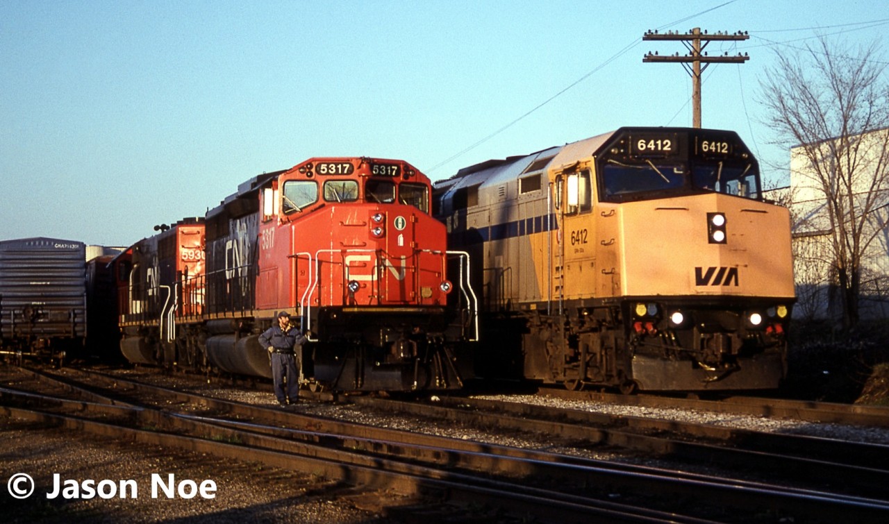 An evening CN 395 is pictured in the yard at Kitchener, Ontario after taking the siding for VIA 89, which is seen passing as it heads to London over the Guelph Subdivision. Powering 395 are SD40-2(W) 5317 and GTW SD40-2 5930, which would soon begin lifting their loaded auto frame cars, once the 15:30 Kitchener Job would clear the yard with GP9RM 7030. The auto frames were manufactured at the massive Budd automotive frame plant in Kitchener situated on the Huron Park Spur.