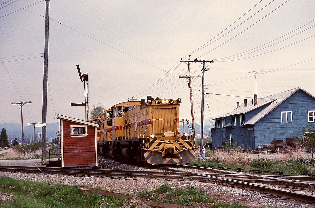 With the Fraser River Bridge at New Westminster out of commission, BC Hydro’s Valley Freight to Huntingdon needed to detour on CP to access interchanges with BN and MILW, resulting in a southward detour crossing the home-road mainline at Clayburn with all three MP15DC units, BCH 152 + 151 + 153, for power on Thursday 1976-04-22.

The little cabin beside CP’s milepost 4 marker enclosed the manual interlocking controls for that diamond crossing.