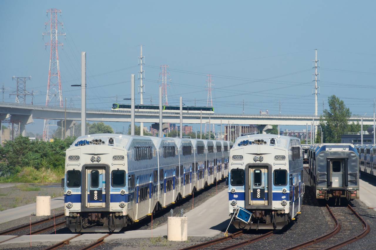 Stored bilevel cars are parked at EXO's maintenance yard in Pointe St-Charles as an REM train passes in the background.